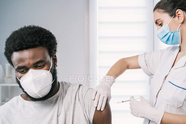 Specialista di medicina femminile in uniforme protettiva, guanti in lattice e maschera facciale che vaccina il paziente afroamericano in clinica durante l'epidemia di coronavirus — Foto stock