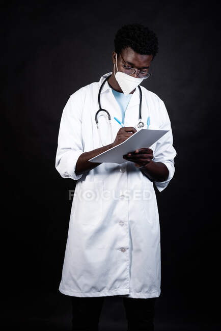 Afroamerikanischer Arzt in Schutzmaske und weißer Uniform macht sich Notizen auf Klemmbrett auf schwarzem Hintergrund — Stockfoto