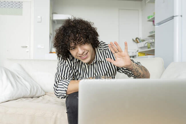 Вміст чоловіка з татуюваннями, що показують привіт жест проти нетбука під час відеочату, сидячи на дивані в квартирі — стокове фото