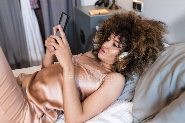 Junge ethnische Frau in Schlafanzügen SMS auf Handy, während sie im Bett im Schlafzimmer liegt — Stockfoto