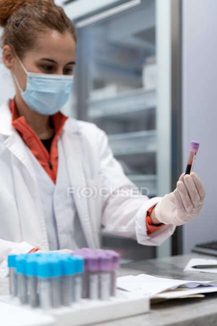 Female medic in mask and gloves holding tube with blood sample in modern lab in clinic — Stock Photo