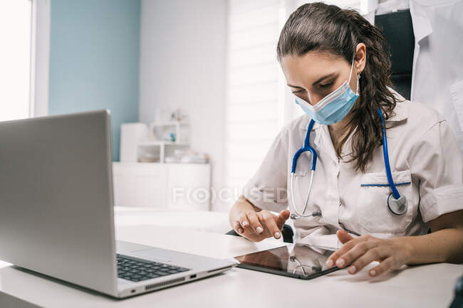 Konzentrierte junge Ärztin in medizinischer Maske und Uniform mit Stethoskop sitzt am Tisch und blättert am Tablet, während sie in modernen Arbeitsräumen arbeitet — Stockfoto