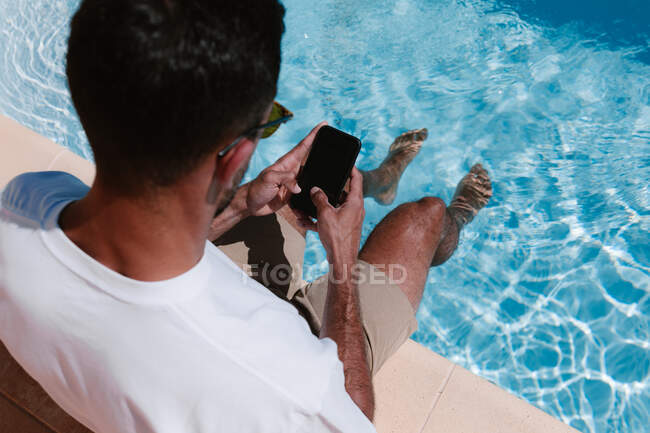 Vista posterior desde arriba de un hombre serio sentado junto a la piscina con las piernas en el agua y navegando en el teléfono móvil durante el trabajo remoto en verano - foto de stock