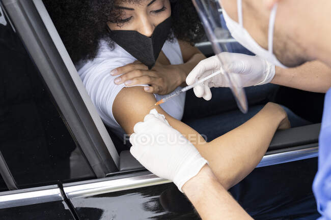 Beschnittene, bis zur Unkenntlichkeit verkleidete Krankenschwester in Latexhandschuhen und Uniform impfende afroamerikanische Patientin im Auto auf einer Fahrt durch eine mobile Klinik während des Coronavirus-Ausbruchs — Stockfoto