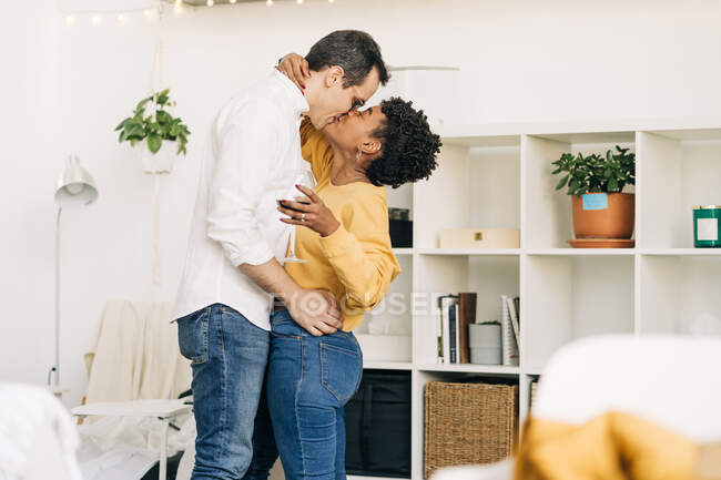 Vista lateral de la sonriente pareja multiétnica enamorada de pie con una copa de vino y besándose mientras pasa un fin de semana romántico en casa - foto de stock
