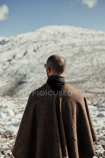 Вид сзади анонимного туриста в мысе, любующегося снежной горой под голубым облачным небом зимой — стоковое фото