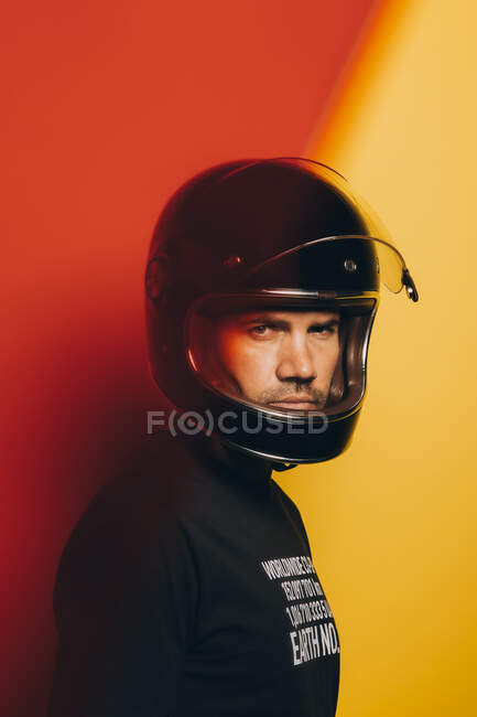 Vista lateral del hombre adulto brutal seguro en casco de motocicleta negro mirando a la cámara mientras está de pie contra el colorido fondo rojo y amarillo - foto de stock