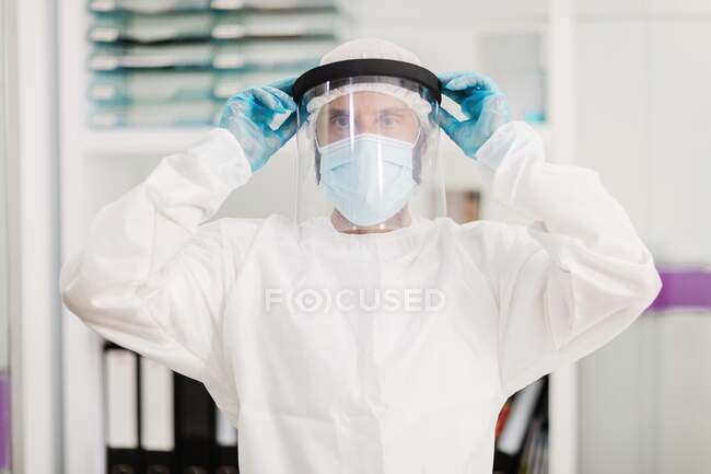 Médico masculino positivo com luvas de látex e uniforme branco colocando máscara protetora de escudo facial em pé no escritório médico moderno e olhando para a câmera — Fotografia de Stock