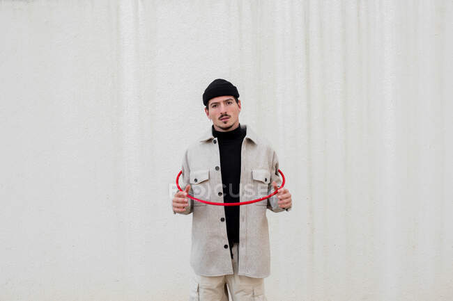 Homme confiant en vêtements à la mode debout avec cerceau de gymnastique rouge contre le mur gris du bâtiment en ville et en regardant la caméra — Photo de stock