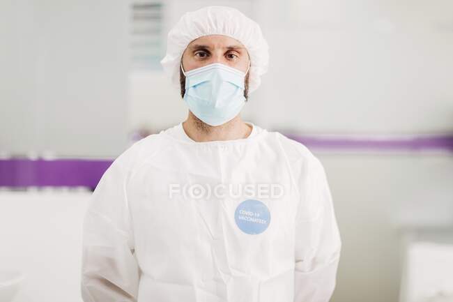 Positiver männlicher Arzt mit Latexhandschuhen und medizinischer Schutzmaske mit Covid-19 geimpften Botschaftsaufkleber auf weißer Uniform steht in moderner Arztpraxis und blickt in die Kamera — Stockfoto