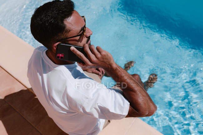 Сверху серьезный мужчина-фрилансер, сидящий у бассейна с ногами в воде и разговаривающий по мобильному телефону во время удаленной работы летом — стоковое фото