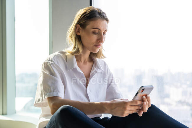 Einsame, emotionslose junge Frau sitzt in leerem Büro mit großem Fenster und surft auf dem Handy — Stockfoto