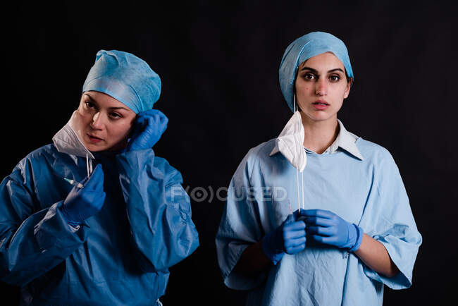 Giovani colleghe in uniforme medica che tolgono maschere facciali mentre sono in piedi su sfondo nero in clinica — Foto stock