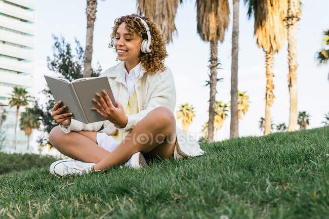 Niedriger Winkel der fröhlichen Afroamerikanerin mit Kopfhörer sitzt im exotischen Park und liest im Sommer interessante Bücher — Stockfoto