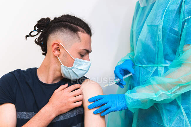 Crop especialista médico fêmea em uniforme de proteção, luvas de látex e máscara facial vacinando paciente hispânico na clínica durante o surto de coronavírus — Fotografia de Stock