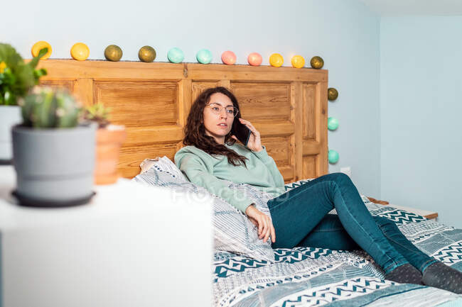 Junge Frau mit türkisfarbenem Sweatshirt und Brille liegt auf dem Bett und telefoniert mit dem Handy — Stockfoto