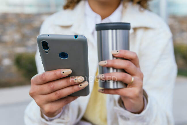 Hembra afroamericana irreconocible recortada con mensajería de café para llevar en las redes sociales a través de teléfonos inteligentes en la calle de la ciudad - foto de stock