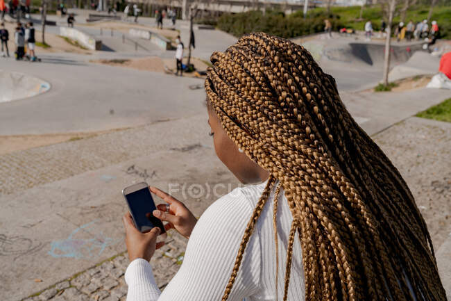 Back view of African American female with braids sitting in skate park and messaging on smartphone on sunny day in summer — Stock Photo