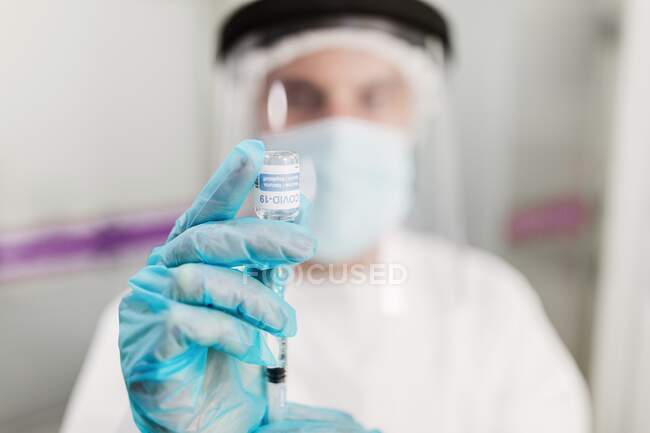 Неопознанный медик в защитной маске и латексных перчатках с флаконом коронавирусной вакцины и шприцем, показываемым на камеру, стоя в больничной палате — стоковое фото