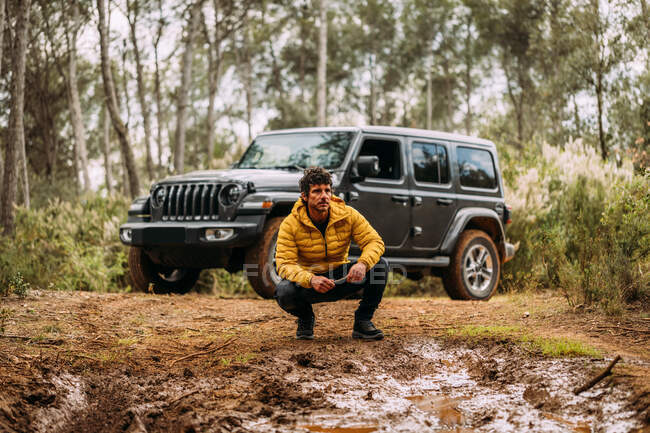 Bottom view of an adventurer crouching next to his off-road car on the mountain — Stock Photo