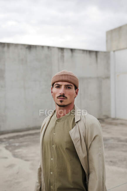 Sério masculino vestindo casaco na moda e chapéu em pé na cidade e olhando para a câmera — Fotografia de Stock