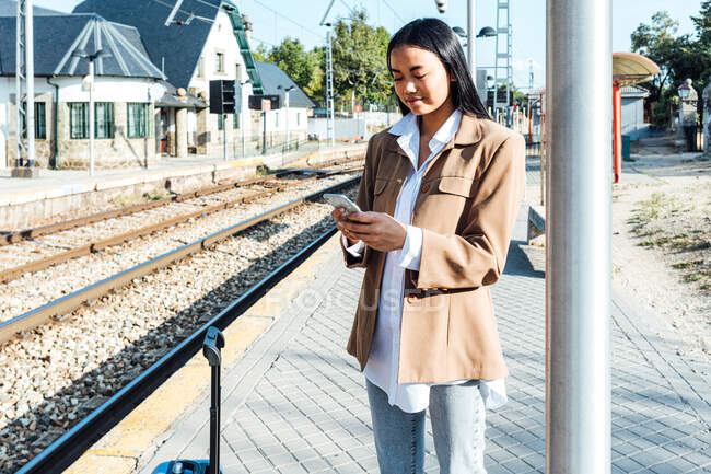 Zufriedene Reisende steht mit Koffer auf Bahnsteig und surft auf Smartphone — Stockfoto