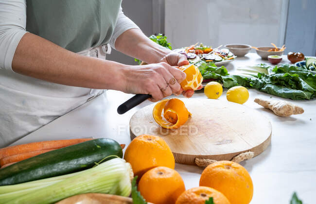 Ernte anonyme Weibchen schälen frische Orange mit Messer über Schneidebrett am Tisch mit Gemüse und Obst — Stockfoto