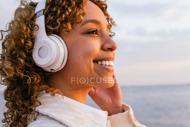 Vista lateral de la alegre hembra negra en auriculares inalámbricos disfrutando de la música en la orilla del mar por la noche - foto de stock