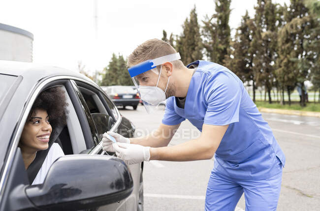 Male doctor in protective uniform, latex gloves and face shield checking African American female patient Identification card before vaccine procedure inside the car on a drive through mobile clinic during coronavirus outbreak — Stock Photo