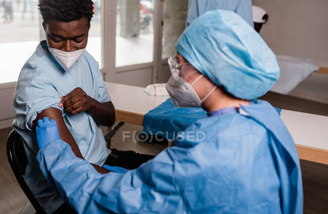 Сторона зору жіночого лікаря в захисній формі і латексні рукавички дезінфікують руку бавовняною тканиною з алкоголем для вакцинації чоловічого афроамериканського пацієнта в клініці під час коронавірусного спалаху. — стокове фото