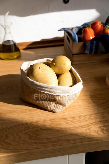Високий кут нагромадження сирої жовтої картоплі в тканинному мішку, розміщеному на дерев'яному столі з інгредієнтами для приготування їжі на домашній кухні — стокове фото
