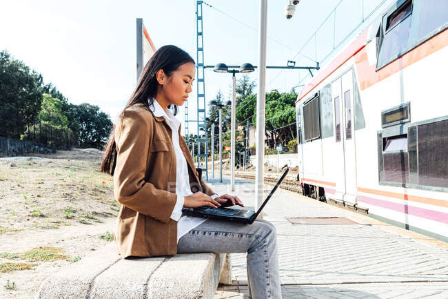 Вид сбоку сосредоточенной азиатской женщины в маске, сидящей на скамейке на вокзале и просматривающей ноутбук в ожидании поезда — стоковое фото
