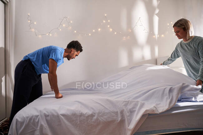 Seitenansicht eines liebenden multiethnischen Paares im Pyjama, das nach dem Aufwachen am Morgen zusammen Bett macht — Stockfoto