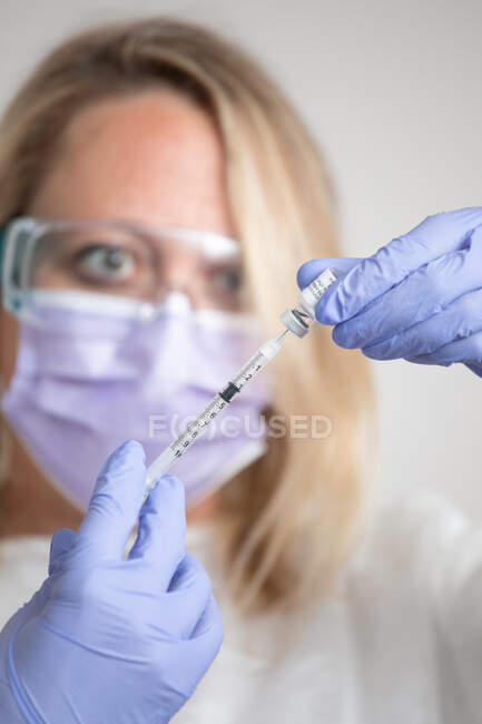 Female medic in protective face shield mask and latex gloves with vial of coronavirus vaccine and syringe showing to camera while standing in hospital room — Stock Photo
