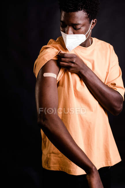 Afro-Américain avec masque protecteur regardant pansement adhésif sur le bras après avoir obtenu la vaccination debout ensemble sur fond noir dans une clinique pendant l'épidémie de coronavirus — Photo de stock