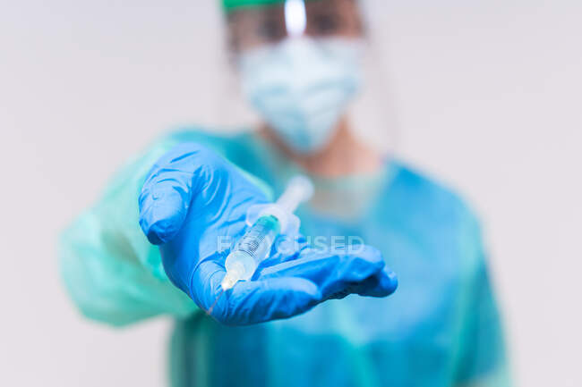Médico borroso irreconocible con máscara protectora y guantes de látex con vial de vacuna contra el coronavirus y jeringa que se muestra a la cámara mientras está de pie en la habitación del hospital - foto de stock