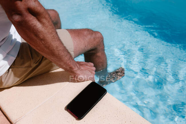 De cima de colheita freelancer masculino irreconhecível sentado à beira da piscina perto de smartphone com tela preta durante o trabalho remoto no verão — Fotografia de Stock