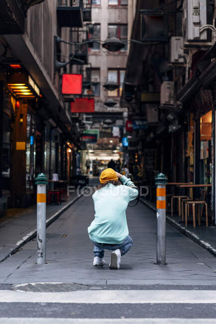 Visão traseira de homem irreconhecível em roupa casual agachando no pavimento enquanto tira foto de edifícios urbanos na câmera — Fotografia de Stock