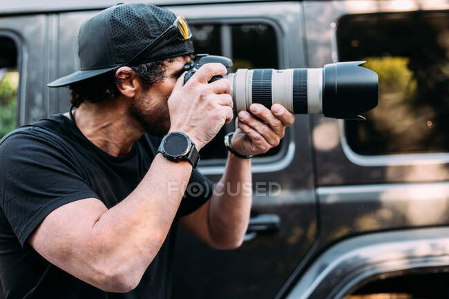 Vista lateral de un fotógrafo aventurero tomando fotos junto a su coche todoterreno - foto de stock