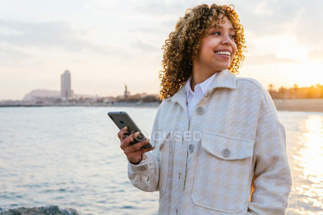 Fröhliche Afroamerikanerin steht am Strand und schaut bei Sonnenuntergang auf dem Smartphone weg — Stockfoto