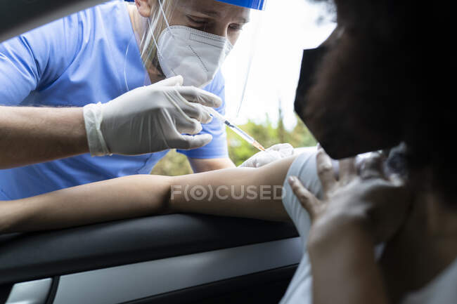 Seitenansicht eines Arztes in Schutzuniform, Latexhandschuhen und Gesichtsschutz, der eine afroamerikanische Patientin während einer Fahrt durch eine mobile Klinik während des Coronavirus-Ausbruchs im Auto impft — Stockfoto