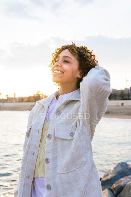 Alto ángulo de alegre hembra afroamericana de pie con los brazos extendidos en las rocas en la orilla del mar y disfrutando de la libertad al atardecer mirando hacia otro lado - foto de stock