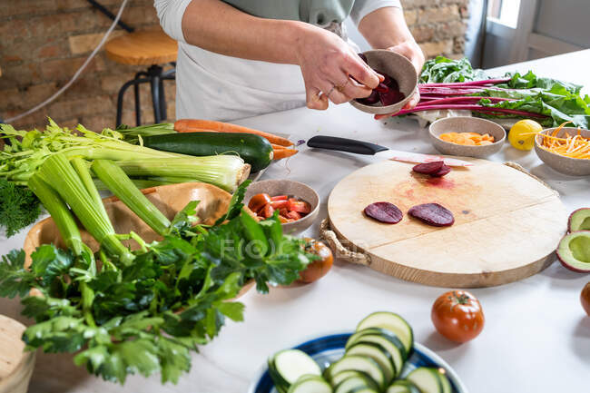 Cultivar hembra irreconocible con remolacha fresca en un tazón con cuchillo mientras se prepara el almuerzo vegetariano en la cocina de la casa - foto de stock