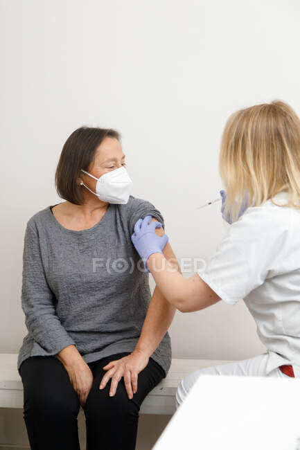 Especialista médica femenina en uniforme protector y guantes de látex vacunando a una paciente mayor en la clínica durante el brote de coronavirus - foto de stock