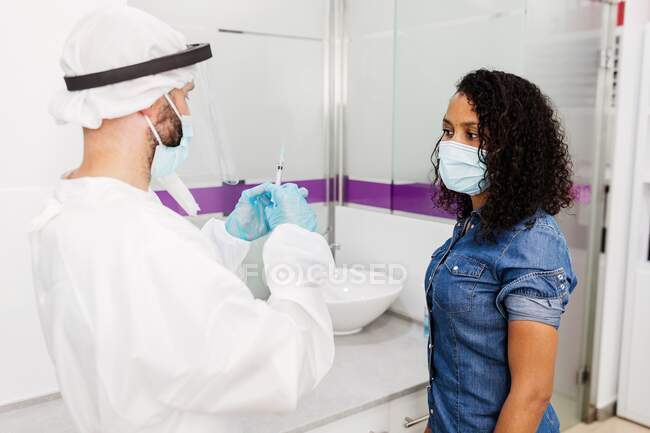 Médico em luvas de látex e protetor facial preenchendo seringa de frasco com vacina preparando-se para vacinar uma paciente afro-americana irreconhecível na clínica durante o surto de coronavírus — Fotografia de Stock