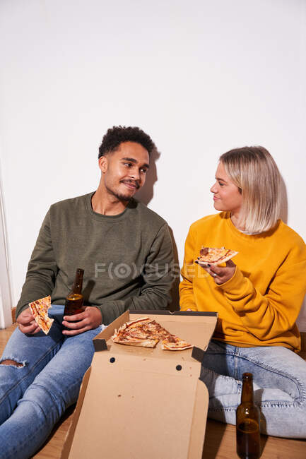 Веселая многонациональная пара, сидящая дома на полу, ест вкусную пиццу и пьет пиво вместе — стоковое фото