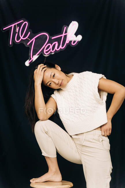 Joven mujer étnica en ropa casual con la pierna en el taburete mirando a la cámara bajo el título - foto de stock