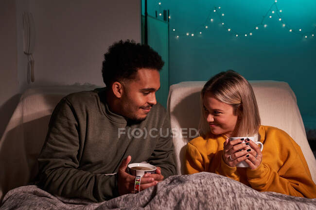 Zufriedenes multiethnisches Paar, das unter einer Decke in Sesseln mit heißen Getränken sitzt und sich beim Genießen des Wochenendes im gemütlichen Wohnzimmer anschaut — Stockfoto