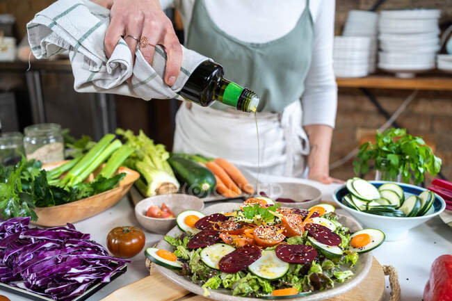 Crop unrecognizable female pouring olive oil from bottle on yummy vegetarian salad with assorted vegetables and sesame seeds at home — Stock Photo