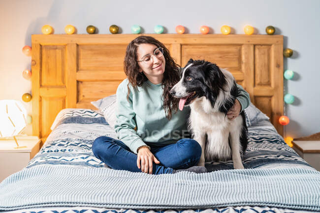 Mujer joven en gafas sentada en la cama con borde collie perro - foto de stock